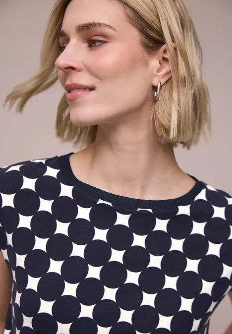 Frau mit kinnlangem blondem Haar, trägt ärmelloses dunkelblaues Top mit weißem Kreisgittermuster.